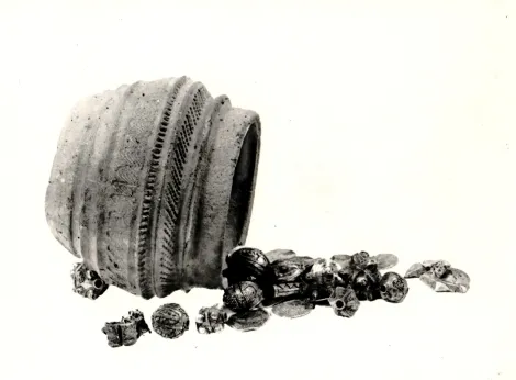 Na zdjęciu widoczne jest przewrócone niewielkie naczynie gliniane, obok niego rozsypane są ozdoby srebrne i monety. Skarb z Ozorkowa w województwie łódzkim, ukryty został w pierwszej ćwierci XI wieku.