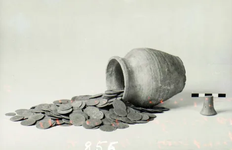 Na zdjęciu widoczne jest naczynie gliniane, obok którego rozsypany jest stos monet. Skarb pochodzi z miejscowości Plęsy w województwie łódzkim i ukryty został około połowy XV wieku.