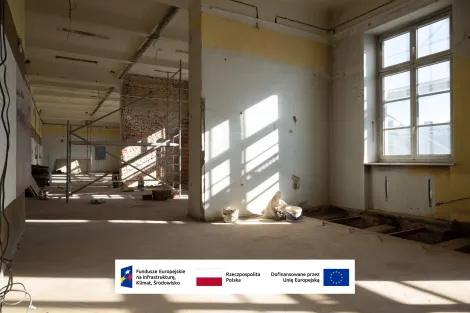 Wnętrze Muzeum Archeologicznego i Etnograficznego w Łodzi. Duże pomieszczenie w trakcie remontu. Odsłonięte ceglane ściany, rusztowanie budowlane, białe wiadra na podłodze i światło słoneczne wpadające przez wysokie okna. Na dole widoczne logotypy Funduszy Europejskich, Rzeczypospolitej Polskiej oraz informacja o dofinansowaniu przez Unię Europejską.