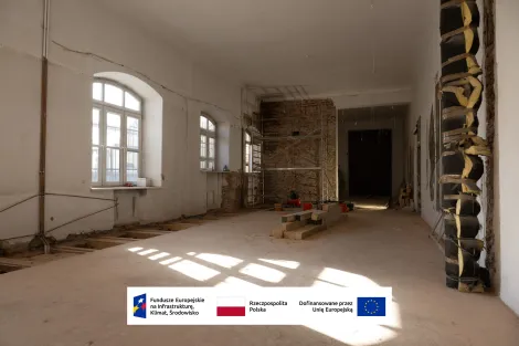 Wnętrze Muzeum Archeologicznego i Etnograficznego w Łodzi. Duże pomieszczenie w trakcie remontu: po lewej stronie trzy wysokie okna wpuszczające światło, odsłonięta ceglana ściana z rusztowaniem na końcu sali, na podłodze ułożone drewniane belki i materiały budowlane, po prawej widoczna izolowana rura. Na dole grafika z logotypami Funduszy Europejskich, Rzeczypospolitej Polskiej i informacją o dofinansowaniu przez Unię Europejską.