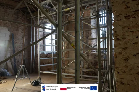Wnętrze Muzeum Archeologicznego i Etnograficznego w Łodzi. Pomieszczenie w trakcie remontu z odsłoniętymi cegłami i fragmentami skutej ściany. Na środku stoi metalowe rusztowanie oraz pionowe drewniane belki konstrukcyjne, pod sufitem widoczne przewody instalacyjne. Na podłodze leży słoma i gruz budowlany, a z lewej strony światło wpada przez wysokie okna. Na dole grafika z logotypami Funduszy Europejskich, Rzeczypospolitej Polskiej oraz informacją o dofinansowaniu przez Unię Europejską.
