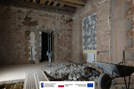 Wnętrze Muzeum Archeologicznego i Etnograficznego w Łodzi. Remontowane pomieszczenie z odsłoniętymi cegłami i fragmentami świeżo zamurowanej ściany. Na podłodze leży sterta gruzu, obok stoi młot wyburzeniowy, a po prawej stronie widoczna jest czarna taczka. W tle znajduje się ciemne przejście do kolejnego pomieszczenia oraz łopata oparta o ścianę. Na dole obrazu widoczny pasek z logotypami Funduszy Europejskich, Rzeczypospolitej Polskiej i Unii Europejskiej.