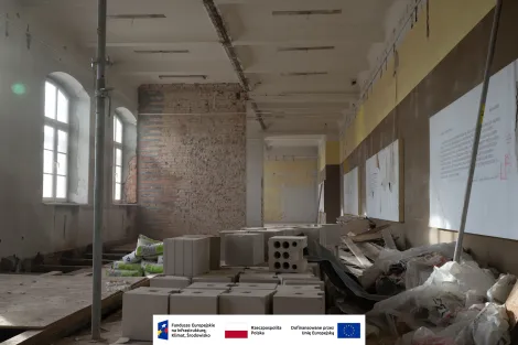 Wnętrze Muzeum Archeologicznego i Etnograficznego w Łodzi. Długie, remontowane pomieszczenie z wysokimi oknami po lewej stronie i częściowo odsłoniętą ceglaną ścianą. Na podłodze oraz prowizorycznych podestach ułożone są białe bloczki budowlane i worki z zaprawą murarską, obok leżą folie oraz deski. Na ścianach wiszą plansze z tekstem, a sufit ma zdemontowane oprawy oświetleniowe i widoczne instalacje. Na dole grafika z logotypami Funduszy Europejskich, Rzeczypospolitej Polskiej i Unii Europejskiej.