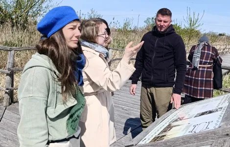 Grupa czterech osób stoi na drewnianej kładce na świeżym powietrzu i ogląda tablicę informacyjną. Dwie kobiety na pierwszym planie  (jedna w niebieskim berecie i zielonej kurtce, druga w jasnym płaszczu i okularach) rozmawiają, a jedna z nich gestykuluje ręką w stronę tablicy. Obok stoi mężczyzna w czarnej bluzie, patrzący na planszę. W tle widać roślinność, drewniane ogrodzenie i bezchmurne niebo. Na dole widoczne logotypy Rzeczypospolitej Polskiej oraz Funduszy Europejskich.