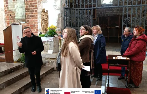 Grupa kilku osób stoi we wnętrzu zabytkowego kościoła z cegły i kamienia. Mężczyzna ubrany na czarno gestykuluje, a pozostali słuchają i patrzą w jego stronę. W tle widać ozdobną metalową bramę, rzeźbę religijną z kwiatami oraz ambonę, a na podłodze leżą czerwone dywany. Na dole widoczne logotypy Rzeczypospolitej Polskiej oraz Funduszy Europejskich.