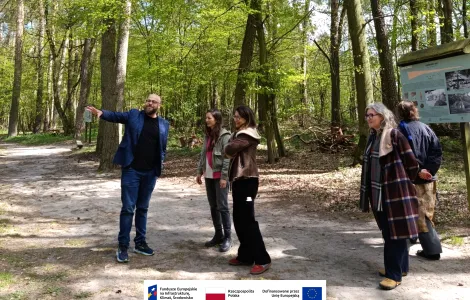 Grupa kilku osób stoi na leśnej ścieżce wśród wysokich drzew. Mężczyzna w niebieskiej marynarce wskazuje ręką w bok, a pozostali uczestnicy spaceru słuchają i patrzą w jego kierunku. W tle widoczna jest tablica informacyjna oraz słoneczna, zielona sceneria lasu. Na dole widoczne logotypy Rzeczypospolitej Polskiej oraz Funduszy Europejskich.