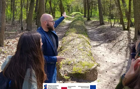 Grupa osób stoi na leśnej ścieżce obok długiego grobowca megalitycznego. Mężczyzna na pierwszym planie gestem ręki wskazuje kierunek wzdłuż nasypu, podczas gdy pozostali słuchają. Wokół rosną wysokie drzewa, a słoneczne światło przebija się przez wiosenne liście. Na dole widoczne logotypy Rzeczypospolitej Polskiej oraz Funduszy Europejskich.