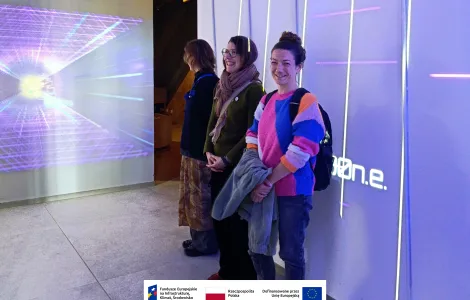 Trzy osoby stoją przy ścianie w nowoczesnej, multimedialnej przestrzeni wystawowej. Na ścianie obok nich widoczna jest projekcja świetlna z geometrycznym wzorem, a pionowe linie świetlne tworzą futurystyczny klimat. Dwie osoby uśmiechają się w stronę aparatu. Na dole widoczne logotypy Rzeczypospolitej Polskiej oraz Funduszy Europejskich.