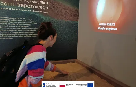 Osoba w muzeum pochyla się nad interaktywną ekspozycją z piaskiem, dotykając jej dłonią. Na ścianie obok wyświetlany jest obraz glinianej amfory z podpisem „Amfora kulista / Globular amphora”. W tle widoczna jest tablica informacyjna dotycząca stanowiska archeologicznego w Brześciu Kujawskim. Na dole widoczne logotypy Rzeczypospolitej Polskiej oraz Funduszy Europejskich.