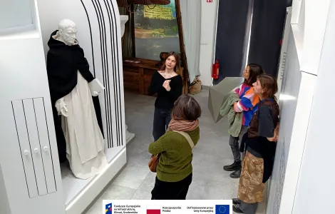 Grupa czterech osób stoi w muzeum przy ekspozycji z figurą w historycznym stroju umieszczoną w podświetlonej wnęce. Jedna osoba, prawdopodobnie przewodniczka, stoi naprzeciw i gestykulując opowiada o eksponacie, podczas gdy pozostali słuchają i obserwują. Wnętrze jest jasne i nowocześnie zaaranżowane, z elementami scenografii nawiązującymi do dawnych czasów. Na dole widoczne logotypy Rzeczypospolitej Polskiej oraz Funduszy Europejskich.