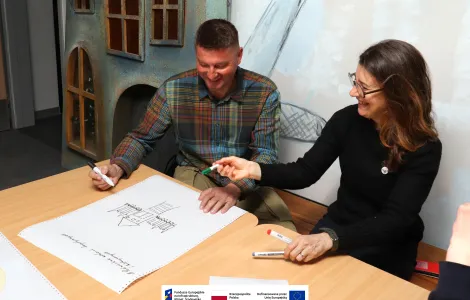 Dwie osoby siedzą przy stole i wspólnie zapisują propozycje na dużej kartce papieru. Mężczyzna w koszuli w kratę trzyma czarny marker i uśmiecha się, patrząc na szkic budynku na kartce. Kobieta obok niego, ubrana w czarny sweter i okulary, podaje mu zielony marker, również uśmiechnięta. Na stole leżą dodatkowe markery i kartki. W tle widać dekoracyjną ścianę z elementami przypominającymi fasadę domu. Na dole widoczne logotypy Rzeczypospolitej Polskiej oraz Funduszy Europejskich.