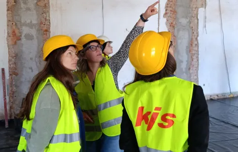 Grupa osób w kaskach ochronnych i odblaskowych kamizelkach stoi wewnątrz remontowanego budynku. Jedna z kobiet wskazuje ręką na fragment ściany, omawiając zakres prac. W tle widać odsłonięte cegły, pęknięcia tynku i przewody elektryczne. Pozostali uczestnicy patrzą w kierunku wskazywanego miejsca i słuchają uważnie. Na dole widoczne logotypy Rzeczypospolitej Polskiej oraz Funduszy Europejskich.