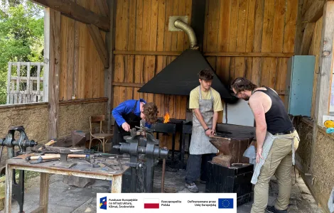 Trzy osoby pracują w kuźni w drewnianej, półotwartej szopie. Na środku pali się ogień w palenisku pod metalowym okapem. Jedna osoba pochyla się nad imadłem, druga trzyma rozgrzany metal przy kowadle, a trzecia obserwuje. Wokół znajdują się narzędzia kowalskie, takie jak młotki, szczypce i kowadło ustawione na solidnej podstawie. Na dole obrazu widoczny jest pasek z logotypami Funduszy Europejskich, Rzeczypospolitej Polskiej i Unii Europejskiej.