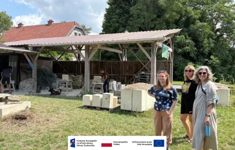 Trzy kobiety stojące na trawie przed zadaszoną, drewnianą wiatą warsztatową. Pod wiatą dwie osoby pracują przy obróbce jasnych, kamiennych bloków, a obok leżą stosy elementów i narzędzia. W tle znajduje się dom z czerwonym dachem oraz drzewa. Na pierwszym planie ustawione są duże, jasne bloki. Na dole obrazu widoczny jest pasek z logotypami Funduszy Europejskich, Rzeczypospolitej Polskiej i Unii Europejskiej.