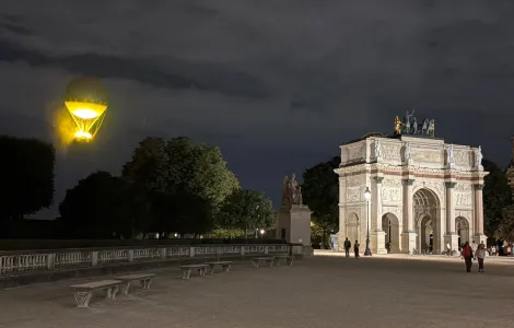 Nocne ujęcie na Łuk Triumfalny na placu Carrousel oświetlonego ciepłym światłem. Na pierwszym planie szeroki, pusty plac z ławkami, w tle kilka spacerujących osób. Nad całością rozciąga się zachmurzone, ciemne niebo. Na dole obrazu znajduje się pasek z logotypami Funduszy Europejskich, Rzeczypospolitej Polskiej i Unii Europejskiej.