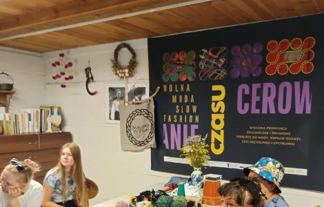 grupa 3 kobiet przy drewnianym stole, przed nimi zestaw materiałów do stworzenia własnej pracy z włóczki, na ścianie baner z napisem "Wolna moda" "Cerowanie czasu"