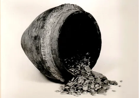 Na zdjęciu widoczne jest przewrócone duże naczynie gliniane, z którego wysypują się placki stopionego srebra, monety i ozdoby. Skarb pochodzi z miejscowości Ruda w województwie łódzkim, ukryty został około połowy XI wieku.