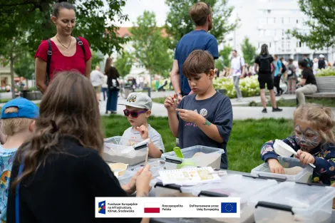 Grupa dzieci podczas warsztatów archeologicznych przy stole piknikowym rozstawionym na zielonej trawie na placu Wolności. W tle tłum osób zebranych na placu. 