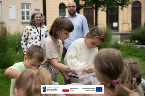 Na pierwszym planie grupa dzieci podczas warsztatów archeologicznych. Na stole stoi plastikowy pojemnik wypełniony piaskiem, wśród którego dzieci próbują odnaleźć ukryte skarby. W tle budynek muzeum oraz kobieta z szarymi włosami w okularach oraz wysoki mężczyzna z brodą w błękitnej koszuli