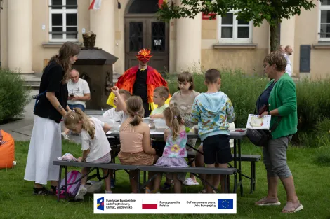 Grupa dzieci podczas warsztatów archeologicznych siedząca przy stole piknikowym. Po lewej animatorka nadzorująca zajęcia, po prawej kobieta w średnim wieku. W tle fontanna miłości z placu Wolności, mężczyzna w średnim wieku, mężczyzna w stroju feniksa oraz budynek muzeum. 