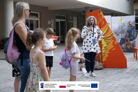 Po prawej stronie kobieta z szarymi włosami w okularach, ubrana w jasną koszulę w brązowe wzory przemawia do mikrofonu podczas otwarcia wystawy. Na prawo od kobiety widoczna rozstawiony fragment mobilnej wystawy plenerowej, która wykonana jest z połączonych ze sobą pionowych elementów (ścianek) utrzymanych w pomarańczowej kolorystyce. Po lewej stronie goście zaproszeni na otwarcie wystawy. W tle budynek na dziedzińcu muzealnym.