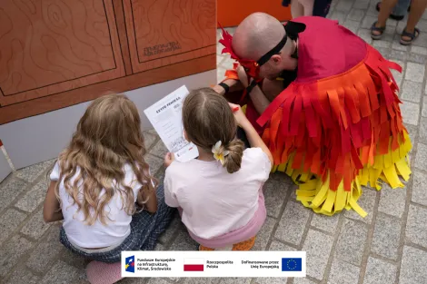 Mężczyzna przebrany za feniksa oraz dwie dziewczynki podczas rozwiązywania quizu 