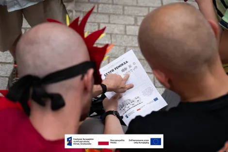 Zbliżenie na kartkę z napisem „Szyfr Feniksa” oraz wskazujący na nią palec. 