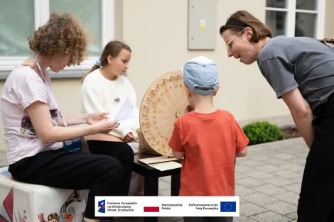 Dwie kobiety oraz dwójka dzieci podczas quizu etnograficznego, na pierwszym planie koło z literami i cyframi wykonane z drewna. 
