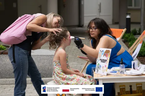 Kobieta wraz z dziewczynką uczesaną w kucyk podczas malowania twarzy. Po prawej malująca z ciemnymi włosami ubrana w kobaltową sukienkę. 