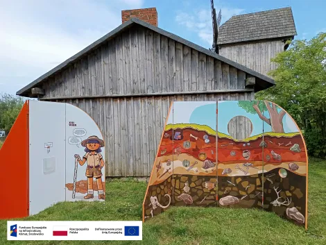 zdjęcie przedstawia ścianki wystawy mobilnej na tle drewnianej zabudowy stodoły. Na jednej ze ścianek widzimy postać archeolożki, na drugiej przekrój warstw ziemi, w której poukrywane są zabytki archeologiczne
