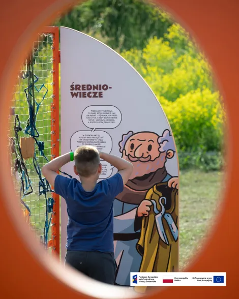 Dziecko stojące tyłem czyta tablicę edukacyjną o średniowieczu. Zdjęcie wykonano przez okrągły otwór w pomarańczowej konstrukcji, który tworzy naturalną ramę kadru. W tle widoczna jest soczysta zieleń roślin i słoneczny dzień. Na dole zdjęcia znajduje się pasek z logotypami Funduszy Europejskich, Rzeczypospolitej Polskiej i Unii Europejskiej.