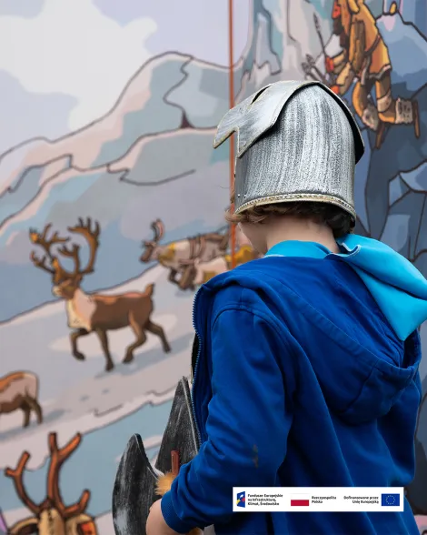 Dziecko w niebieskiej bluzie i stylizowanym hełmie ogląda tablicę edukacyjną z ilustracjami reniferów i górskiego krajobrazu. Na dole zdjęcia znajduje się pasek z logotypami Funduszy Europejskich, Rzeczypospolitej Polskiej i Unii Europejskiej.
