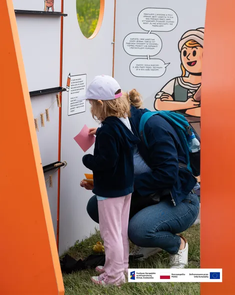 Dziecko w jasnej czapce i różowych spodniach stoi przy interaktywnym panelu edukacyjnym na świeżym powietrzu, trzymając kolorowy arkusz papieru, obok klęczy dorosła osoba w dżinsach i granatowej kurtce. Panel jest biało-pomarańczowy, z rysunkową postacią kobiety w historycznym stroju. . Na dole zdjęcia znajduje się pasek z logotypami Funduszy Europejskich, Rzeczypospolitej Polskiej i Unii Europejskiej.