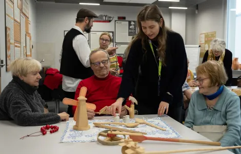 Przy stole siedzi troje starszych ludzi. Między nimi stoi młoda kobieta, a z tyłu jeszcze stoi stoi dwoje ludzi. Na stole leżą drewniane zabawki.
