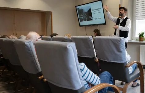 Mężczyzna w białej koszuli i czapce stoi z podniesioną ręką w górę. Obok widać ekran a na nim zdjęcie. Przed nim na szarych fotelach siedzą ludzie.