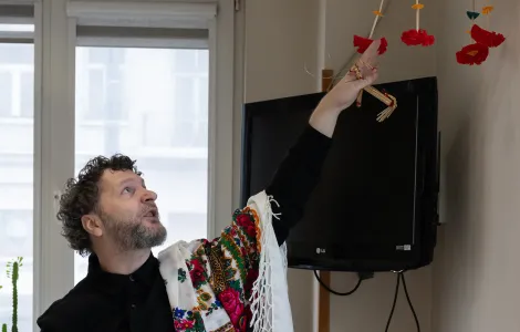 Mężczyzna w czarnej bluzce i chustce w kwiaty zarzuconej na ramię, pokazuje palcem na wiszący kolorowy pająk.