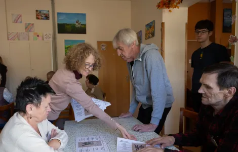Kobieta w średnim wieku pokazuje ręką na plansze, które leżą na stole. Przy stole siedzą kobieta i mężczyzna, a obok nich stoi starszy mężczyzna.