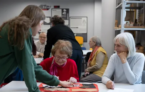 Dwie starsze kobiety siedzą przy stole i oglądają matę. Mata jest czerwona z naszytym futerkiem. Młoda kobieta pokazuje im coś na macie.