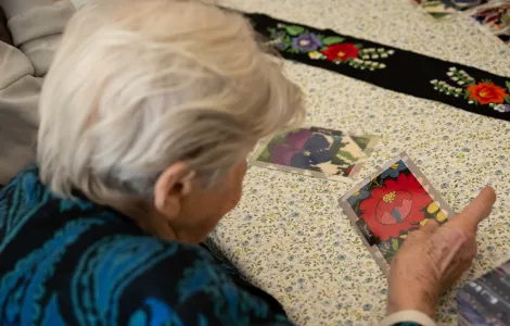 Starsza siwa pani siedzi przy stole i ogląda zdjęcie wyhaftowanej róży. Na stole leży zdjęcie innego haftu oraz pas z wyhaftowanymi kwiatami.