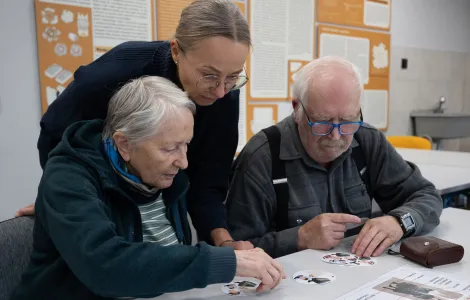 Przy stole siedzi starsza kobieta i starszy mężczyzna. Między nimi stoi kobieta w średnim wieku. Wszyscy oglądają kółka z kolorowymi obrazkami. 