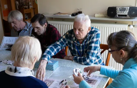 Trzech mężczyzn i dwie kobiety siedzą przy stole. Ludzie są starsi. Oglądają kolorowe kółeczka i plansze.