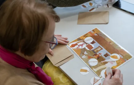 Starsza kobieta siedzi przy stole. Na stole lezy obrazek pokazujący wiejską chałupę. Na obrazku są ułożone krążki z obrazkami.